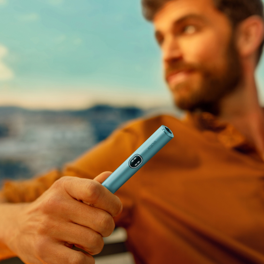 Baiat brunet cu bluza portocalie care tine in mana un stilou IQOS iluma i