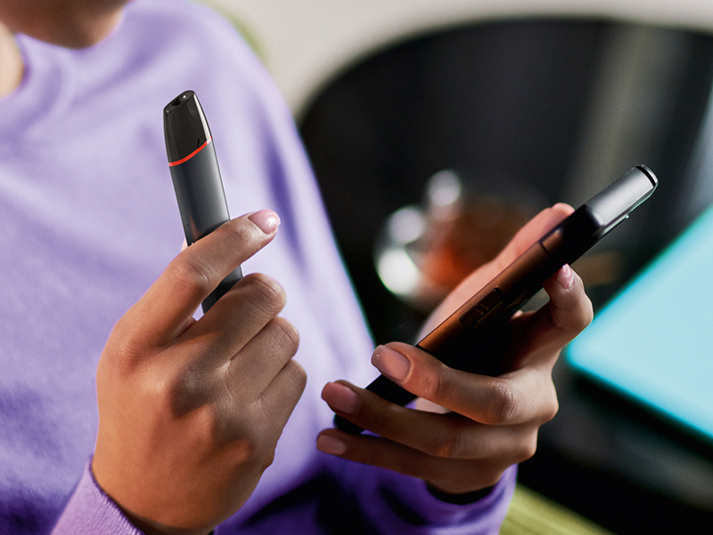 Imagine - Mâini care țin un dispozitiv de vapat VEEV inPRIME negru și un telefon