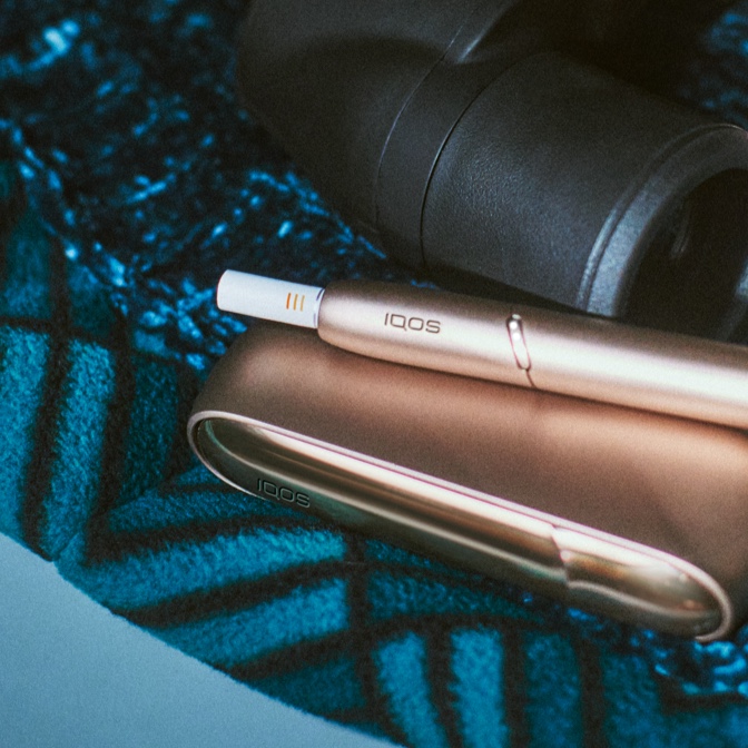 Încărcător IQOS gold pe un material albastru, alături de un binoclu